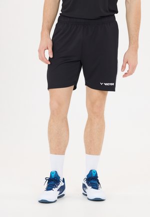 Mand iført sorte Victor sportsshorts, hvide høje sokker og hvide-blå sports sko, stående mod en ensfarvet baggrund.