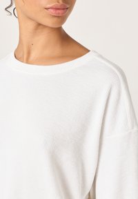 Kvinne iført en hvit ribbestrikket genser med rund hals, som viser nakke, kjeve og en del av skulderen.