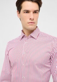 Camicia bianca da uomo con sottili righe verticali rosse, colletto classico e pattina frontale con bottoni bianchi. Tessuto leggermente lucido.