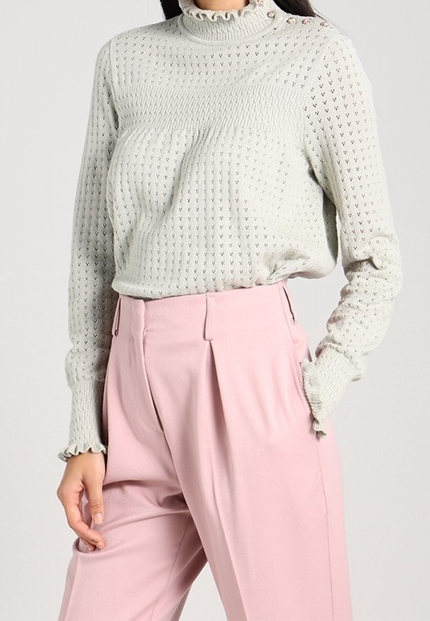 Pull en tricot gris clair avec un col haut à volants et des accents de boutons, associé à un pantalon tailleur rose pâle avec des plis.