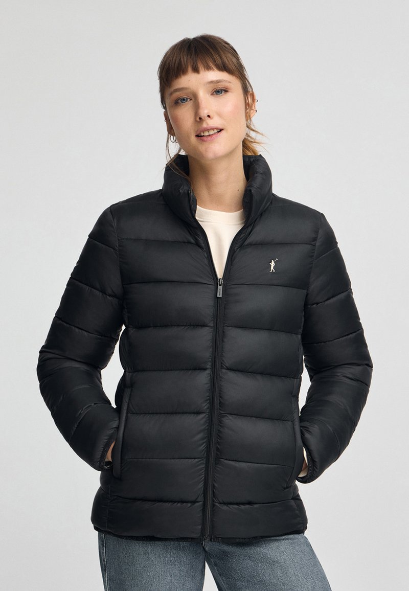Svart puffjacka med vertikal kviltad design, hel dragkedja och hög krage. Har en liten logotyp på vänster bröst.