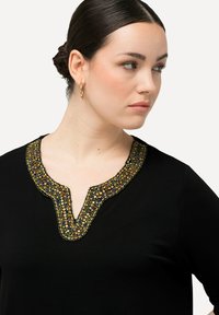 Top nero con scollatura a V adornato con perline colorate in tonalità di oro, blu e verde. Tessuto morbido con maniche corte.