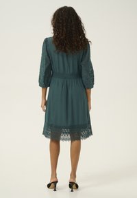 Abito teal con maniche in pizzo a lunghezza gomito, vita smockata e lavorazione in pizzo intricata all'orlo. Tessuto liscio con una vestibilità leggermente ampia.