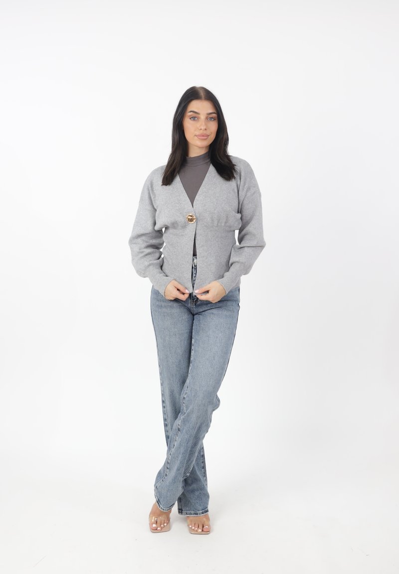 Cardigan grigio con chiusura a bottone, realizzato in un tessuto morbido. Indossato sopra una maglietta scura, abbinato a jeans chiari e sandali trasparenti.