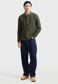 Olivengrüne Strickjacke mit Kragen und Knopfverschluss, kombiniert mit dunkelblauen, locker geschnittenen Jeans und beigen Schuhen.