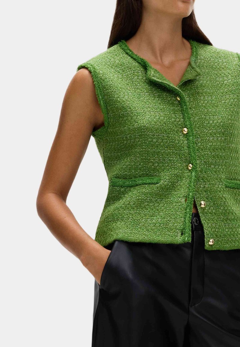 Gilet verde breve senza maniche realizzato in un tessuto testurizzato, caratterizzato da un colletto, bottoni dorati e due tasche frontali. Indossato sopra pantaloni neri.