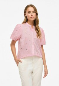 Blouse rose pâle à manches courtes avec un col rond, ornée de broderies florales et de motifs découpés, associée à un pantalon de couleur crème.