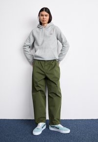 Grauer Kapuzenpullover mit Fronttasche, grüne weit geschnittene Hose und blaue Turnschuhe mit weißen Sohlen, vor einem schlichten Hintergrund.
