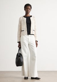 Donna che indossa un cardigan a maglia crema sopra una maglietta nera, pantaloni bianchi a gamba larga, scarpe nere e porta una borsa nera, in piedi su uno sfondo neutro.