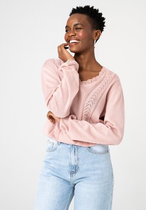 Kobieta uśmiechająca się, z krótkimi kręconymi włosami, ubrana w różowy dzianinowy sweter z koronkowymi detalami oraz jasnoniebieskie dżinsy, na białym tle.