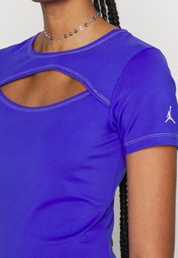 Top ajustado morado con cuello redondeado, una abertura en forma de ojo de cerradura en el pecho, mangas cortas y costuras blancas contrastantes. Logo de Jumpman en la manga.