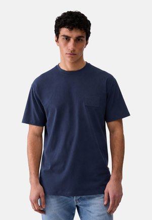Uomo con capelli corti e ricci che indossa una t-shirt blu scuro tinta unita a maniche corte e jeans azzurri su sfondo bianco.