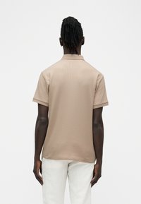 Homme à la peau foncée et aux dreadlocks portant un polo à manches courtes côtelé beige et un pantalon blanc, debout de dos sur un fond blanc.