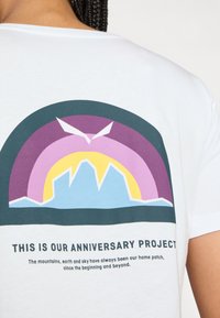 Weißes T-Shirt mit einem bunten grafischen Design von Bergen und einem Regenbogen, mit dem darunter stehenden Text "DAS IST UNSER JUBILÄUMSPROJEKT."