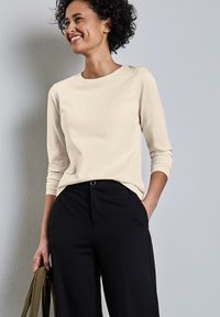 Beige pitkähihaisessa paidassa on pyöreä kaula-aukko, ja se on yhdistetty mustiin leveisiin housuihin. Kangas vaikuttaa sileältä ja kevyeltä.