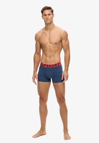Marineblauwe katoenen boxershorts met een elastische tailleband met "SUPERDRY" in het rood. Model staat op blote voeten en toont een slimfit ontwerp.