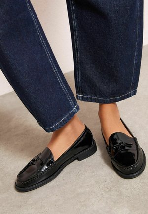 Füße in glänzenden schwarzen Leder-Loafers mit Quasten, kombiniert mit dunkelblauen, gerade geschnittenen Jeans auf einem neutralen Boden.