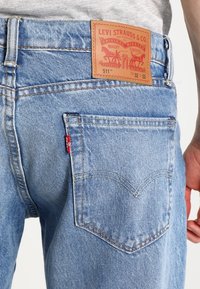 Jeans de mezclilla azul claro con un ajuste de tiro medio, que presentan una etiqueta de cuero con branding en rojo y dorado en la cintura y bolsillos traseros con costura.