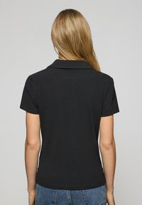 Svart polo skjorta med korta ärmar och krage, med en slät tygstruktur och en figurnära design. Bakifrån syns rena linjer och sömmar.