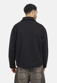 Veste en polaire noire avec un col haut, conçue avec une coupe décontractée. Présente des manches longues et une texture douce. La vue de dos montre peu de détails.