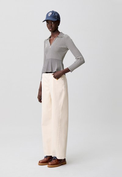 Top de manga larga a rayas, pantalones anchos color crema y zapatos marrones con cordones. La modelo lleva una gorra azul oscuro con emblema. Atuendo sencillo y casual.