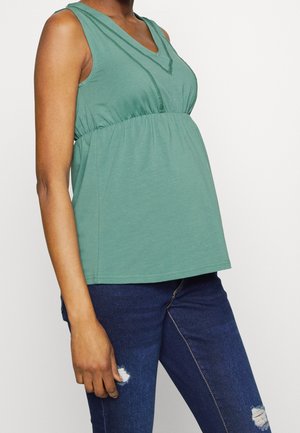 Personne enceinte portant un haut sans manches vert et un jean déchiré bleu foncé, se tenant devant un fond clair uni.