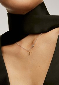 Collar de cadena de plata con un colgante en forma de letra "E" dorada, adornado con brillantes acentos y un pequeño ornamento redondo, luciendo sobre un fondo negro.