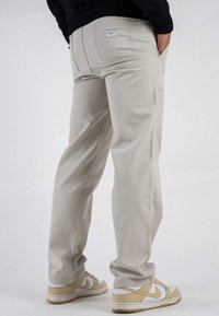 Pantaloni cargo beige con tasche posteriori, taglio dritto e una texture liscia. Abbinati a sneakers beige e bianche, rivolti leggermente di lato.