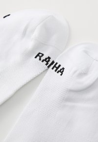 Weiße Sportsocken mit einem strukturierten Strickmuster, versehen mit schwarzer "RAHA"-Beflockung an der Seite. Der Zehenbereich hat eine glatte Oberfläche.