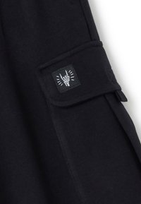 Schwarzer Stoff mit einer Seitentasche, die ein schwarzes Patch mit einem weißen Handgeste-Design zeigt. Glatte Textur mit sauberer Naht.