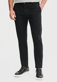 Czarne spodnie wykonane z gładkiego, elastycznego materiału, o standardowym kroju, z zapięciem na guzik i bocznymi kieszeniami, noszone z czarnymi trampkami.