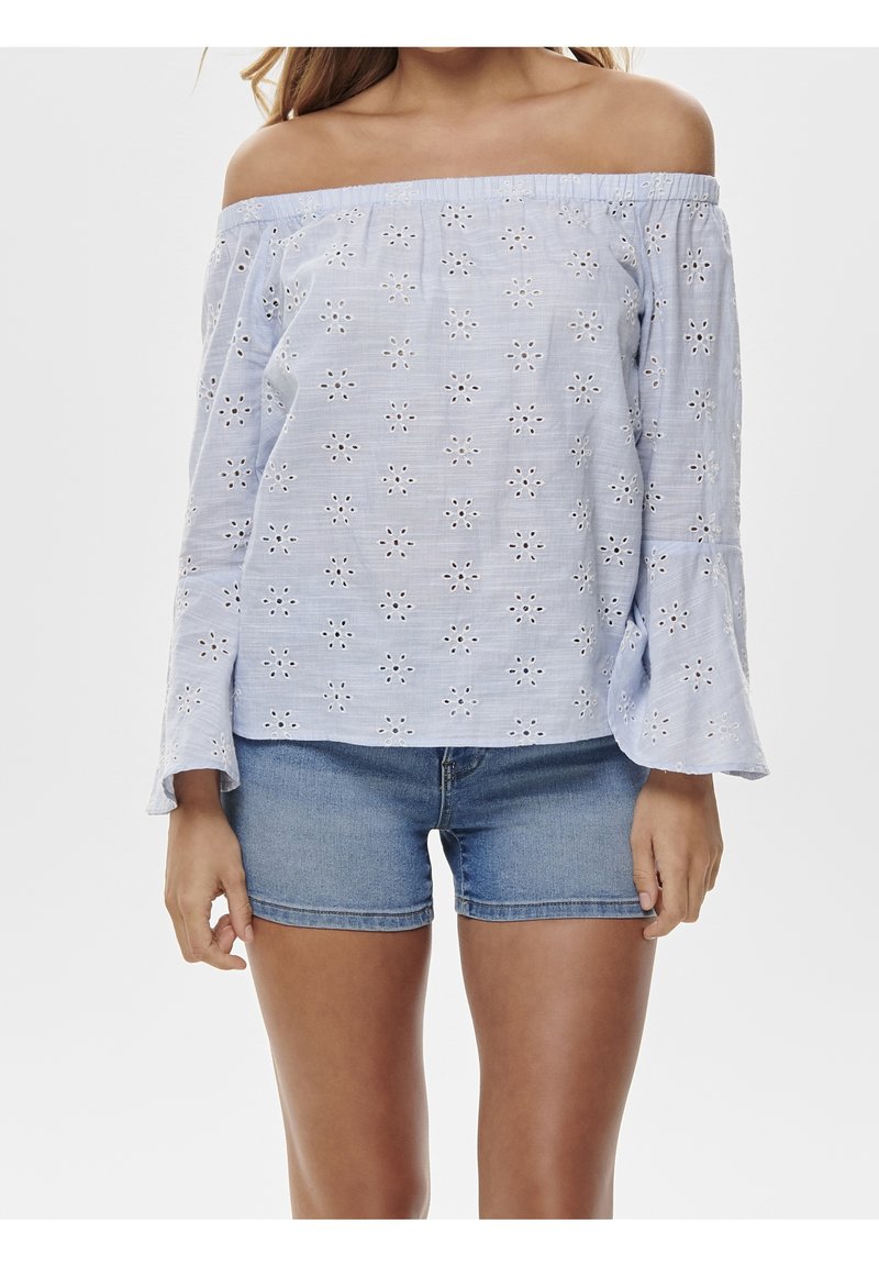 Blusa azzurro chiaro con spalle scoperte e ricami floreali bianchi, maniche a campana e una vestibilità rilassata. Abbinata a pantaloni corti di denim.