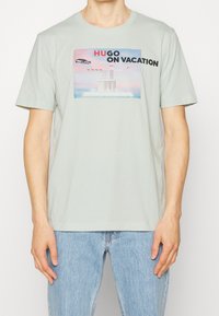 Camiseta de algodón verde claro con un estampado gráfico que incluye el texto "HUGO ON VACATION", mostrando un coche y un edificio contra un fondo de atardecer.