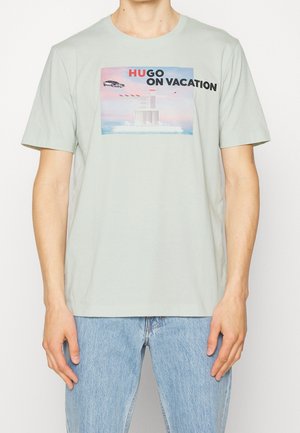 T-shirt en coton vert clair avec un motif graphique comportant le texte "HUGO EN VACANCES", représentant une voiture et un bâtiment sur fond de coucher de soleil.