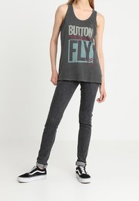 Camiseta de tirantes gris con el texto "BUTTON FLY", combinada con jeans oscuros y zapatillas en blanco y negro. Textura suave, ajuste casual, estilo relajado.