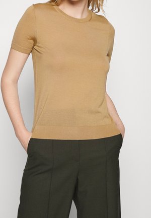 Kobieta nosząca dopasowany krótki sweter w kolorze camel wsunięty w wysokie spodnie w ciemnym oliwkowym odcieniu, z rękami w kieszeniach.