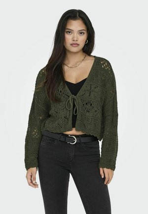 Cardigan corto verde oliva con un motivo di crochet testurizzato, dettagli con laccetto e maniche lunghe. Indossato sopra un top nero aderente e abbinato a jeans scuri.
