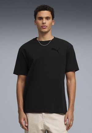 Joven con camiseta negra de Puma, pantalones beige, collar de cadena plateada y anillos, de pie frente a un fondo de degradado gris.