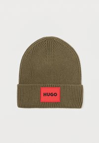 Căciulă tricotată de culoare verde măslin, cu un manșon pliat, având un eticheta rectangulară roșie cu cuvântul "HUGO" scris cu litere negre.