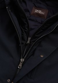 Laivastonsininen takki, jossa on kaksinkertaiset vetoketjut ja sileä pinta. Vuori on pehmustettu. Mukana on ruskea kangasetiketti, jossa on teksti "OSCAR JACOBSON".