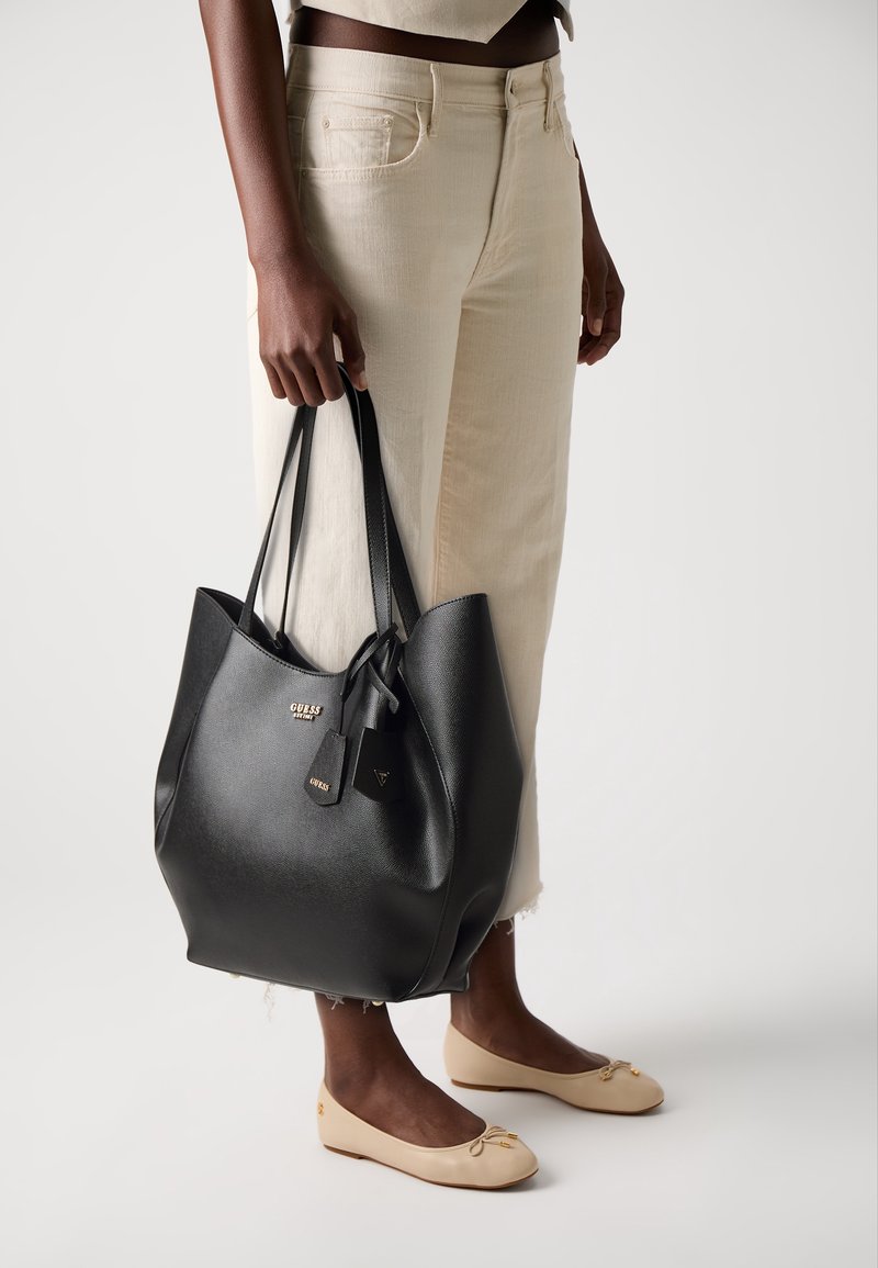 Personne portant un jean court beige clair et des ballerines beiges tenant un grand sac cabas en cuir noir Guess avec double poignées.