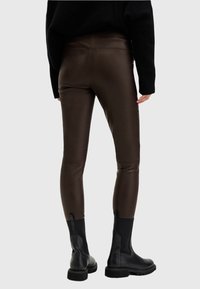 Osoba w ciemno-brązowych dopasowanych skórzanych spodniach i czarnych botkach na kostkę, stojąca na tle jasnym.