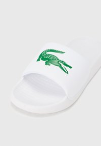 Λευκή σαγιονάρα με υφή στο πέλμα και φαρδιά λουράκι που φέρει λογότυπο πράσινου κροκόδειλου. Λείο υλικό με μινιμαλιστικό σχεδιασμό.