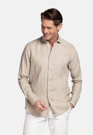 Man glimlachend, een beige overhemd met lange mouwen aan het dichtknopen, draagt witte broek, staand tegen een effen lichte achtergrond.