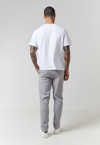 Hvit bomulls t-skjorte parret med lysegrå bukser, begge med avslappet passform. Modell som har på seg hvite joggesko, vist fra baksiden.
