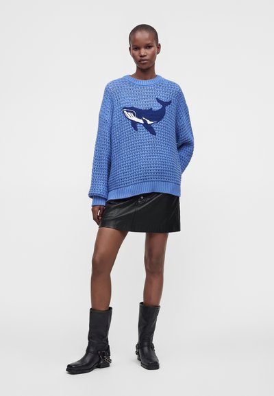 Femme portant un pull en maille bleu avec un motif de baleine, une mini-jupe en cuir noire et des bottes noires mi-mollet, debout devant un fond uni.