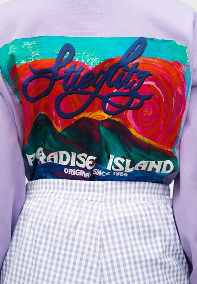 Πουκάμισο λεβάντας με πολύχρωμο γραφικό σχέδιο βουνών και ήλιου, κείμενο "Stiegltz Paradise Island Original Since 1988", συνδυασμένο με μπλε φούστα με καρό σχέδιο.