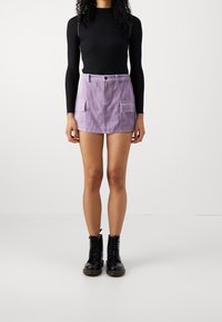 Fialová kordurová sukně s dvěma předními kapsami, v kombinaci s černým topem s dlouhým rukávem a černými šněrovacími botami. Sukně má zapínání na knoflík.