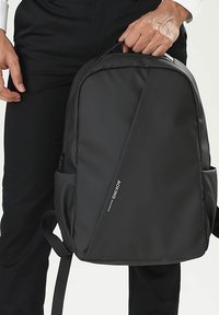 Sac à dos noir en matériau lisse et durable. Présente un design structuré et épuré avec une poche latérale zippée et des bretelles réglables.