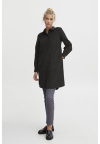 Čierna tunika s golierom, dlhými rukávmi, prednými gombíkmi a vreckom na hrudi, skombinovaná so šedými slim-fit nohavicami a čiernymi loafermi.